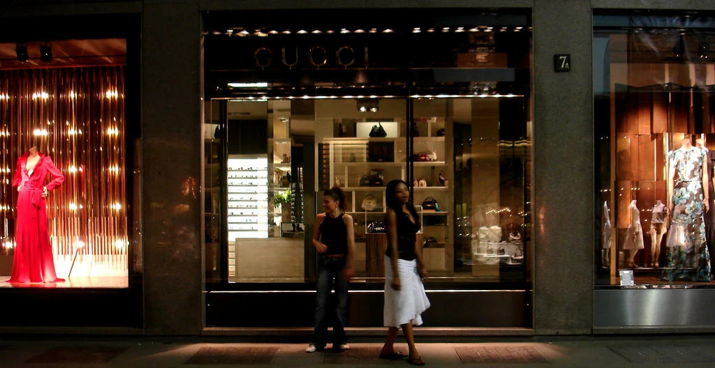 Gucci storefront on Via Montenapoleone in Milan at night, with illuminated window displays and pedestrians outside.