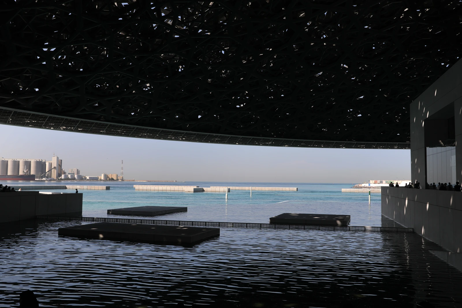 Louvre Abu Dhabi beneath its latticed dome, with reflecting pools and the waterfront visible beyond.