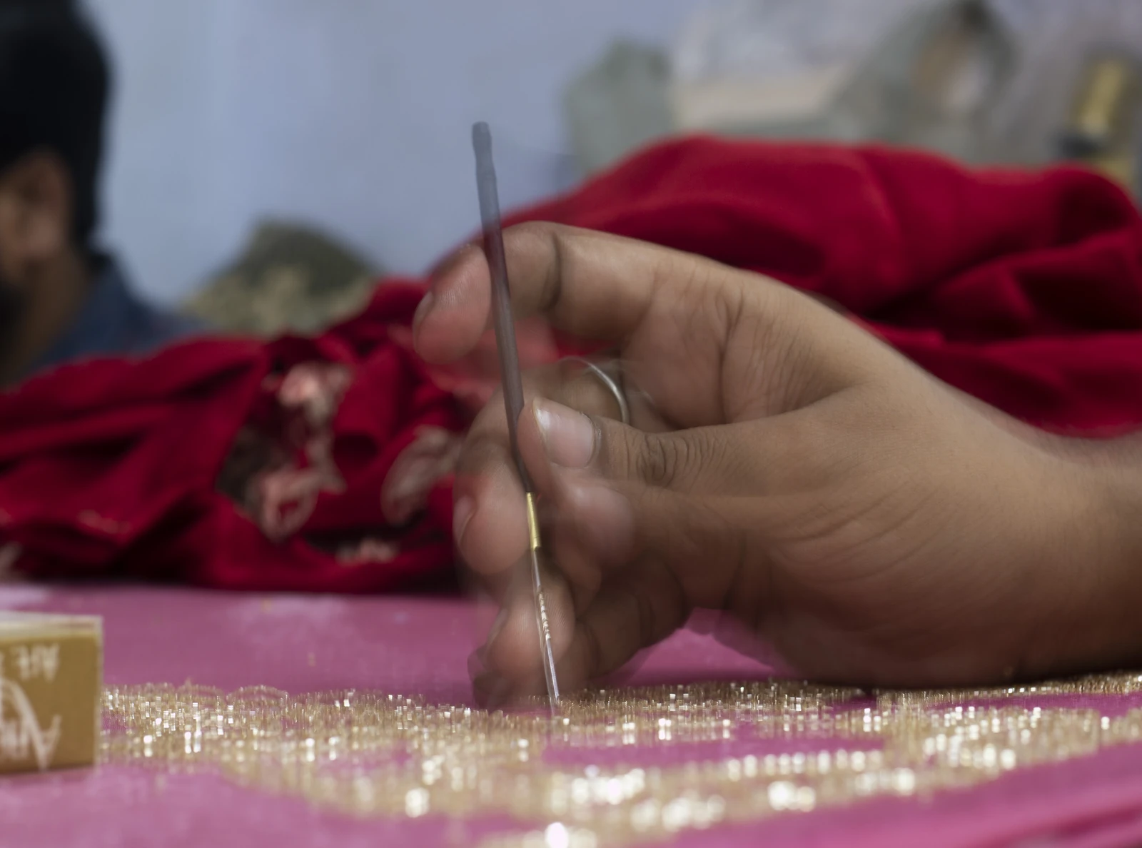 Artisan hand stitching metallic zardozi embroidery on red fabric in India.
