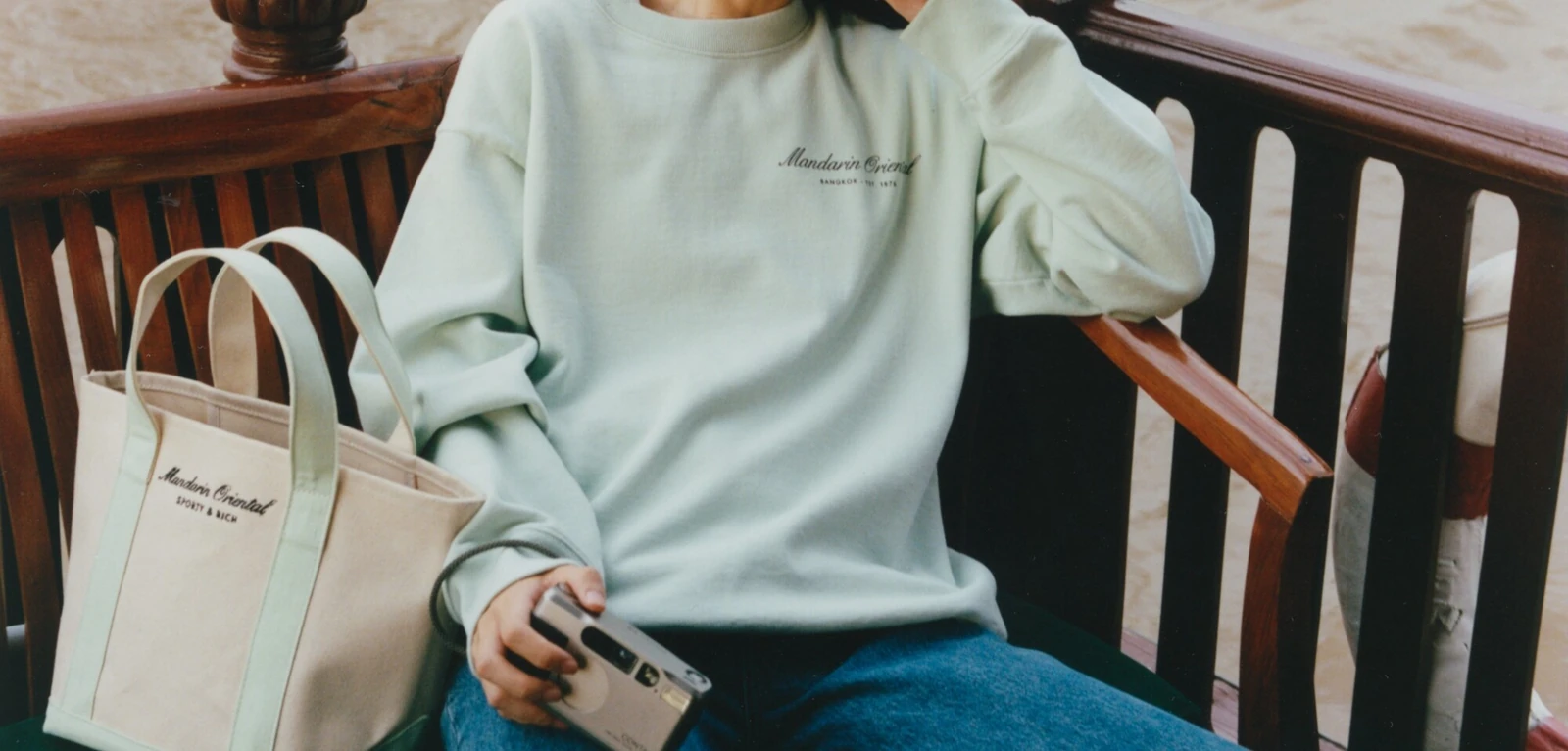 Person wearing a pale green Mandarin Oriental Bangkok x Sporty & Rich sweatshirt beside a matching canvas tote bag.
