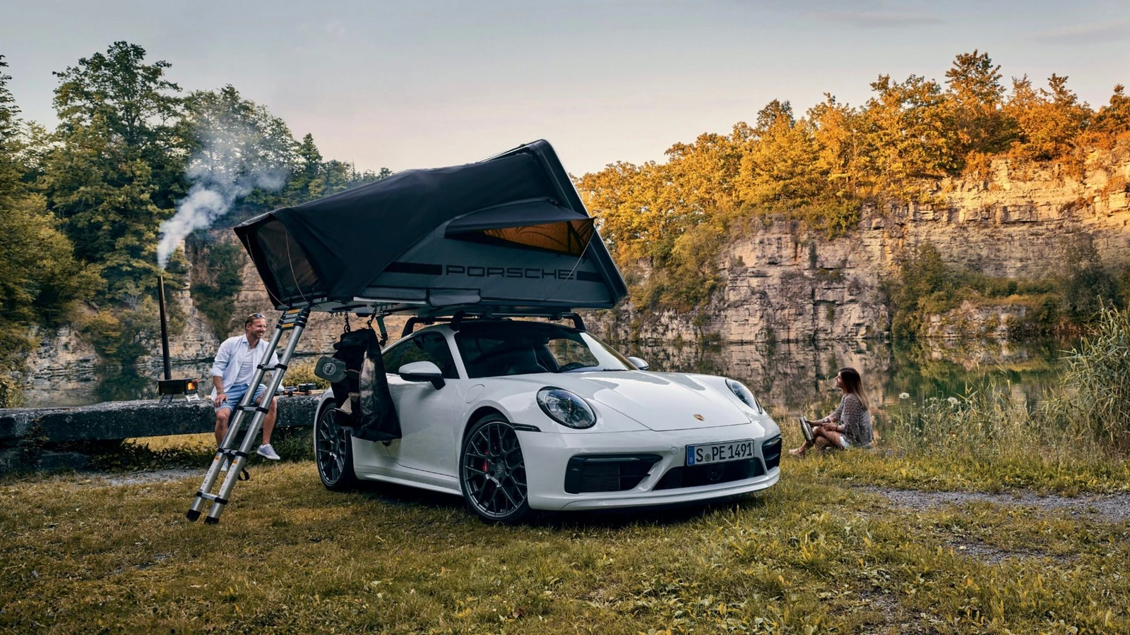 A white Porsche sports car beside a rocky lakeshore with the black Porsche Tequipment roof tent deployed on its roof.