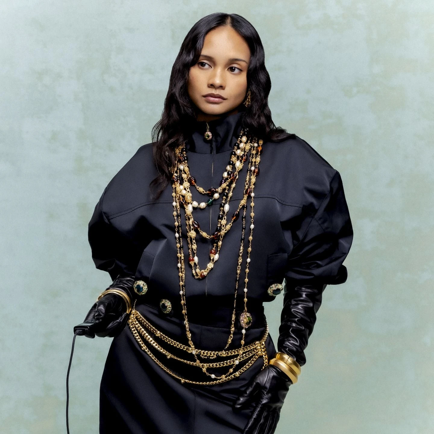 Model wearing a black St. John Fall 2026 look with layered gold-and-pearl necklaces, chain belts and black gloves.
