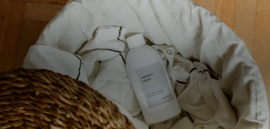 Unbranded laundry detergent bottle in a basket with white linens on a wooden floor.