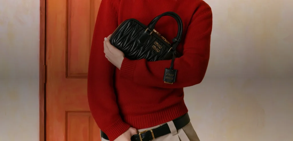 Gigi Hadid in a red sweater holding a black quilted Miu Miu Arcadie handbag in front of an orange door.