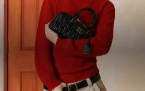 Gigi Hadid in a red sweater holding a black quilted Miu Miu Arcadie handbag in front of an orange door.