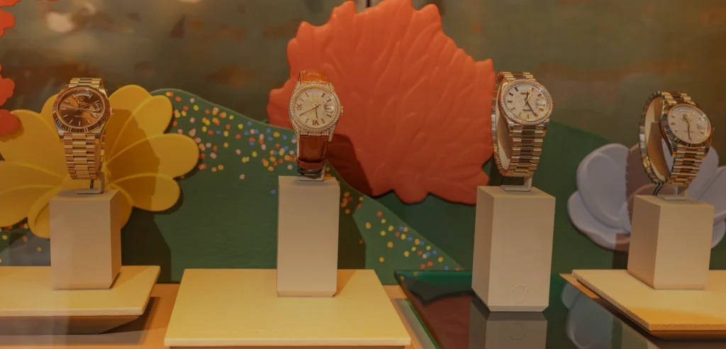 Four Rolex watches displayed on cream stands against a colorful boutique window backdrop.